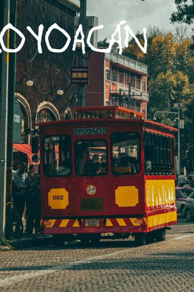 A vibrant red and yellow trolley travels along the streets of Coyoacán in Mexico CIty, showcasing its colorful design against the urban backdrop.